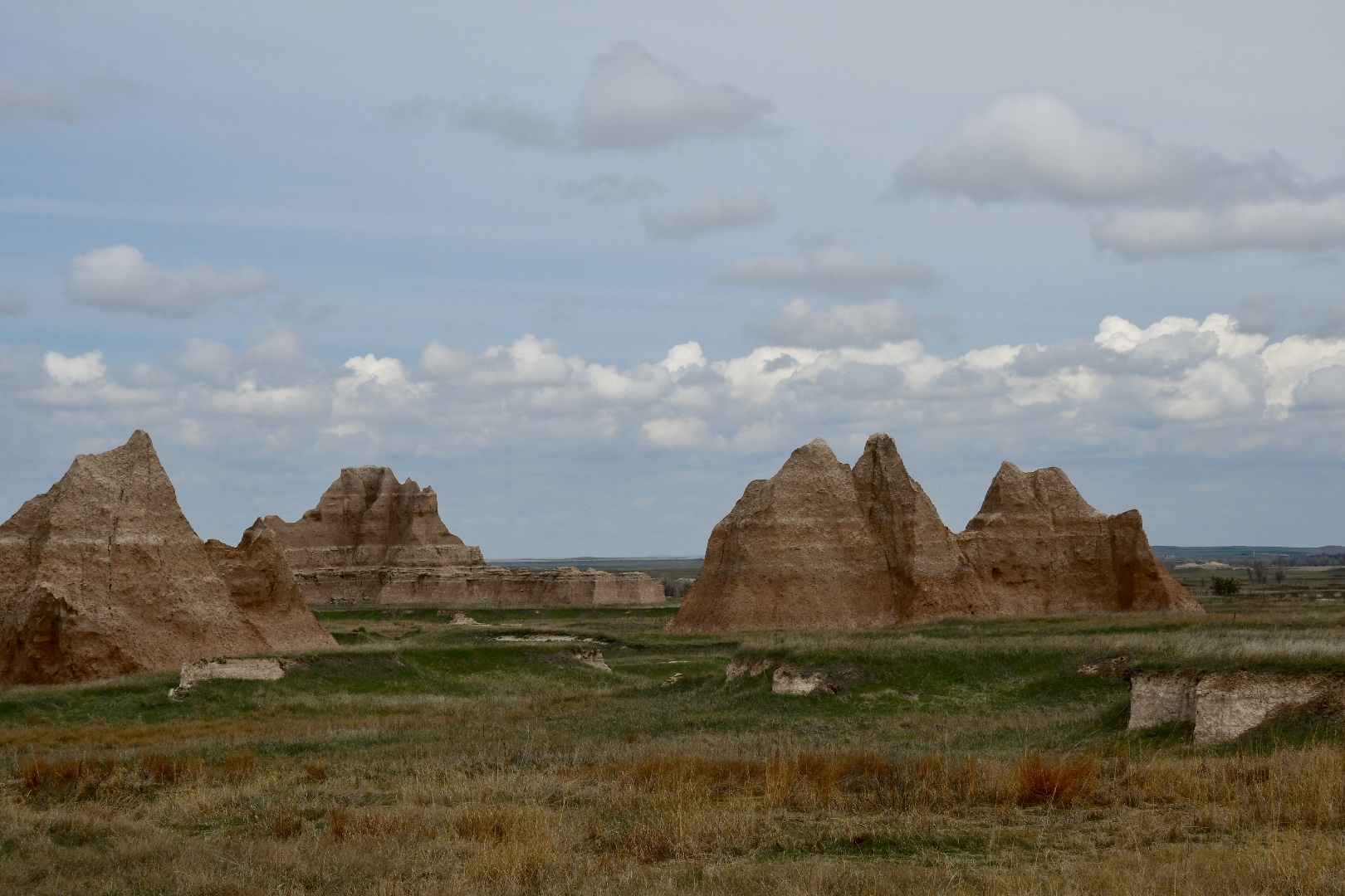 Im Badlands National Park ragen aus dem grünen Gras rote Felsen in dreieckiger Tipiform in den blauen Himmel mit weißen Wolken