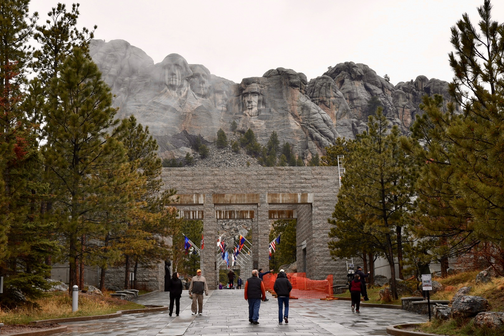 Besucher auf einem gepflasterten Spazierweg zum Mount Rushmore, ein von Nadelbäumen eingerahmtes Steinportal ist der Durchlass zum Weg mit vielen Fahnen